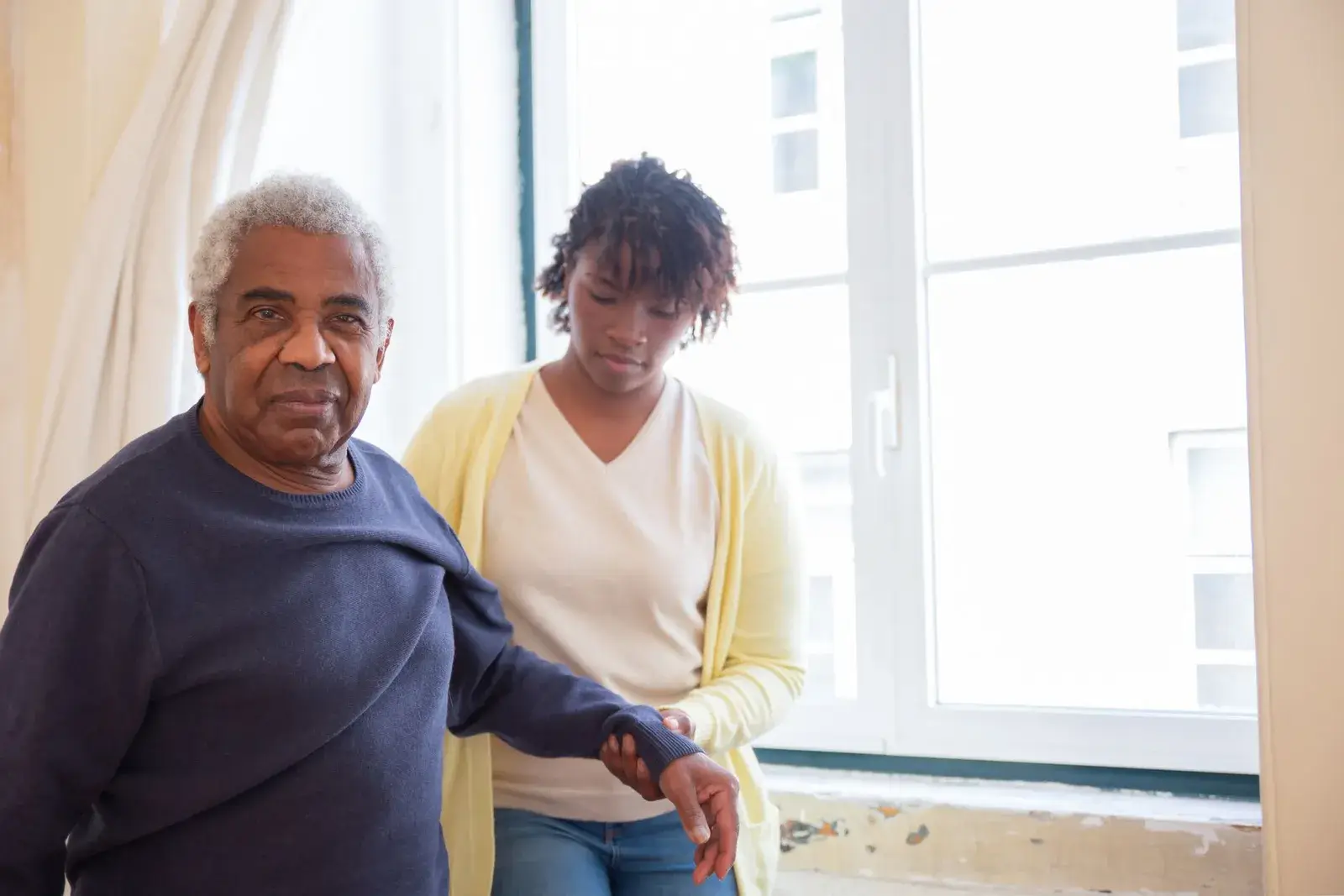 A clinician supporting an older man by the arm as he walks beside a bright window